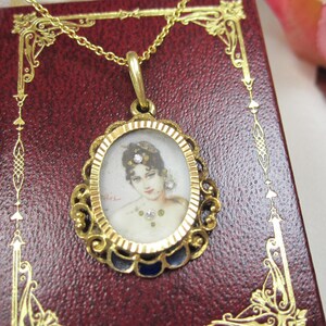 May include: A gold-toned pendant necklace featuring an oval miniature portrait of a woman. The portrait is set within a decorative frame with blue enamel accents. The necklace rests on a red leather-bound surface.