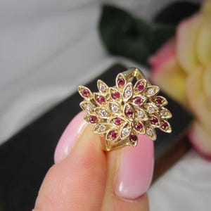 May include: A gold ring featuring a cluster of marquise-shaped stones arranged in a leaf-like pattern. The design alternates between deep red stones and clear, sparkling white stones, all set within a polished yellow gold metal band structure.