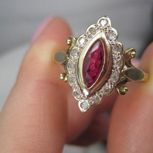 May include: A gold ring featuring a central marquise-cut deep red gemstone set in a bezel, surrounded by a halo of small round clear stones. The setting has an ornate, elongated shape with decorative metal accents on the sides of the band.
