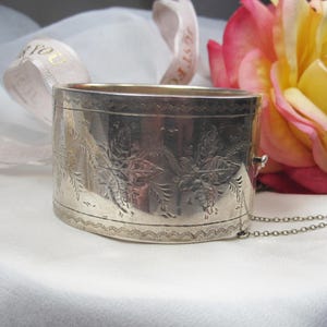 Op de afbeelding: Een antieke zilverkleurige armband, ovaal van vorm, met bloemengravures. Het heeft een scharniersluiting en een fijne ketting. Een roze en gele roos en een wit lint met gouden tekst staan op de achtergrond.