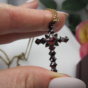 Peut inclure: Un collier doré avec un pendentif en forme de croix. La croix est ornée de pierres précieuses rouge foncé, avec une pierre carrée centrale et de petites pierres facettées le long des bras. La chaîne est délicate, et le pendentif est tenu par une main.