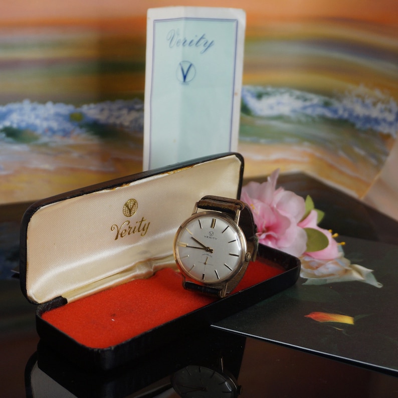 Op de afbeelding: Een goudkleurig Verity-polshorloge met een bruine leren band ligt in een open zwarte koffer met een rode binnenkant. De wijzerplaat van het horloge is zilverkleurig met gouden wijzers. Een Verity-brochure staat erachter.