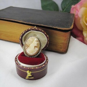 May include: An oval shell cameo brooch featuring a carved profile of a person, surrounded by a delicate border of tiny seed pearls. The brooch is displayed inside a small, round, burgundy-colored velvet-lined jewelry box with a gold-tone latch.