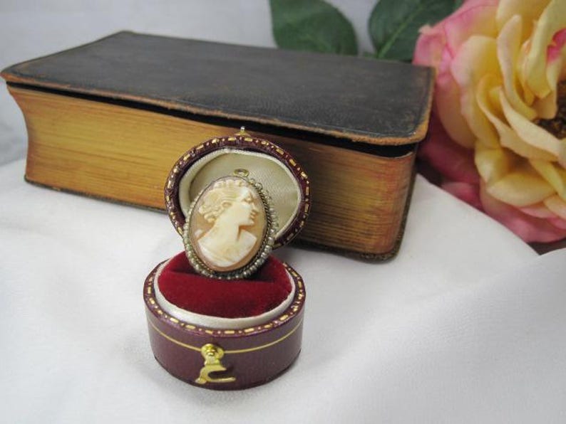 May include: An oval shell cameo brooch featuring a carved profile of a person, surrounded by a delicate border of tiny seed pearls. The brooch is displayed inside a small, round, burgundy-colored velvet-lined jewelry box with a gold-tone latch.