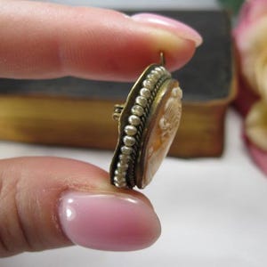 May include: An oval-shaped cameo brooch featuring a carved profile relief set in a brass-toned metal frame. The edge of the frame is decorated with a row of small white seed pearls and a twisted metal wire border, with a small metal loop at the top.
