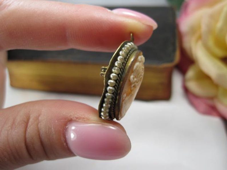 May include: An oval-shaped cameo brooch featuring a carved profile relief set in a brass-toned metal frame. The edge of the frame is decorated with a row of small white seed pearls and a twisted metal wire border, with a small metal loop at the top.