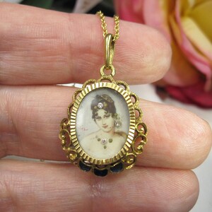 May include: A gold-toned pendant necklace featuring an oval portrait of a woman. The portrait is framed by an ornate gold filigree design with a dark blue enamel border. The necklace has a delicate gold chain and a small loop at the top.