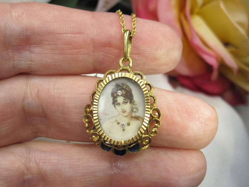 May include: A gold-toned pendant necklace featuring an oval portrait of a woman. The portrait is framed by an ornate gold filigree design with a dark blue enamel border. The necklace has a delicate gold chain and a small loop at the top.