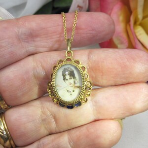 May include: A gold-toned pendant necklace featuring an oval portrait of a woman. The pendant has an ornate filigree frame with blue enamel accents. The chain is delicate, and the necklace is held in a hand.