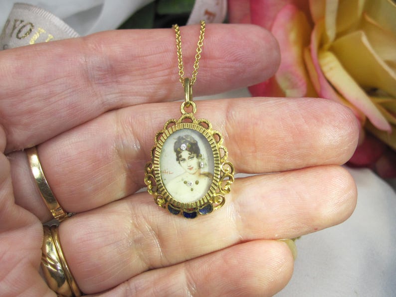 May include: A gold-toned pendant necklace featuring an oval portrait of a woman. The pendant has an ornate filigree frame with blue enamel accents. The chain is delicate, and the necklace is held in a hand.