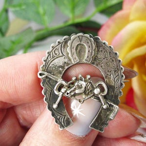 Puede incluir: Broche vintage plateado con diseño de herradura y ecuestre. El broche presenta una herradura en la parte superior, una cabeza de caballo con riendas y detalles florales. El broche está sujeto por una mano.