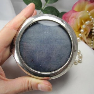 May include: A round, silver-toned metal compact case featuring a smooth, polished frame and a circular, dark blue fabric-covered center panel. The object has a reflective, metallic border and a simple, circular design with a flat, textured surface.