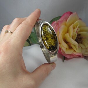 May include: A silver hinged bracelet featuring an oval amber stone. The amber stone is a translucent yellow-green colour with inclusions. The bracelet is held by a hand, with a gold ring on the finger. A pink and yellow rose is in the background.