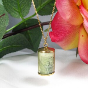 May include: A gold pendant necklace with a cylindrical locket. Inside the locket is a rolled-up paper, possibly a banknote. The necklace is set against a backdrop of a rose and green leaves.