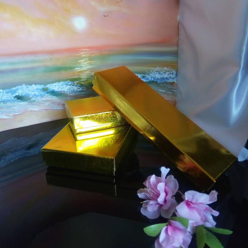 May include: Three gold boxes stacked on top of each other. The boxes are shiny and have a textured surface. The boxes are in front of a painting of a sunset over the ocean.