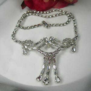 Può includere: Una collana color argento con strass trasparenti e scintillanti. La collana presenta un design a doppio filo, con un pendente centrale di strass a cascata e pendenti a goccia. Il gioiello è esposto su una superficie bianca.