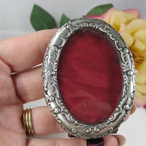 May include: An oval-shaped silver frame featuring an intricate embossed scrollwork pattern along the border. The center of the frame is filled with a deep red fabric inlay. Small hallmark stamps are visible on the left side of the silver frame.