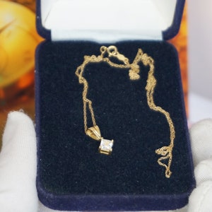 May include: A gold chain necklace with a square-shaped pendant featuring a clear gemstone. The necklace is displayed in a blue velvet jewellery box.