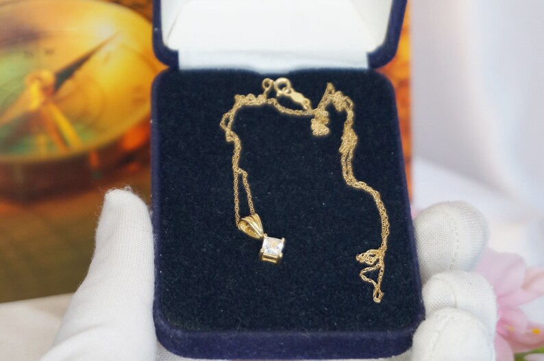 May include: A gold chain necklace with a square-shaped pendant featuring a clear gemstone. The necklace is displayed in a blue velvet jewellery box.