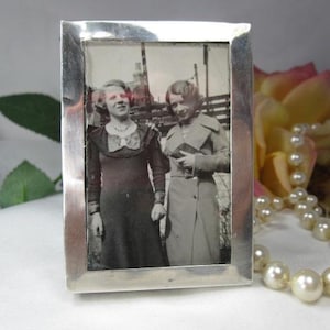 May include: A rectangular silver picture frame containing a black and white photograph of two individuals standing side by side. The frame has a polished, reflective metallic finish and a simple, clean border surrounding the vintage portrait.