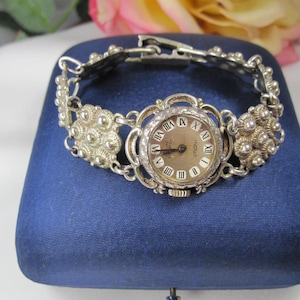 May include: Silver-toned vintage watch with a round face and Roman numeral hour markers. The watch features a decorative band with floral-like accents. The watch is displayed on a blue velvet surface, with a rose and greenery in the background.