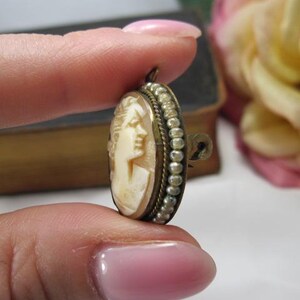 May include: An oval-shaped cameo brooch featuring a carved cream-colored profile of a person. The central carving is framed by a border of small, round, white seed pearls set in a dark metal casing with a twisted wire edge and a pin mechanism.