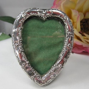 May include: A heart-shaped silver picture frame with an ornate, scrolling design. The frame has a green velvet backing. A yellow and pink flower is in the background.
