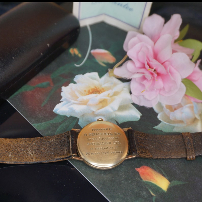 Op de afbeelding: Een goudkleurig polshorloge met een bruine leren band. De wijzerplaat van het horloge is zichtbaar en de achterkant van het horloge heeft gegraveerde tekst. Het horloge wordt getoond met roze en witte bloemen en een zwarte etui.