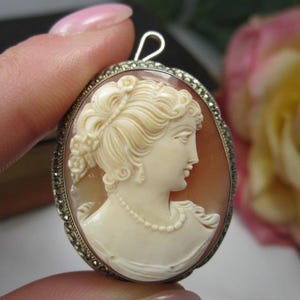 May include: An oval cameo pendant featuring a cream-colored carved profile of a person with an elaborate updo hairstyle and a beaded necklace. The cameo is set in a silver-toned metal frame decorated with small, dark, faceted stones and a bail.
