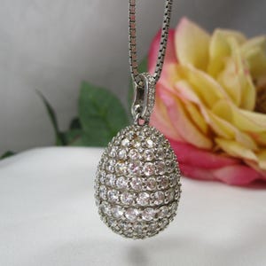 Pode incluir: Um colar com pingente de prata com um pingente redondo em forma de ovo incrustado com numerosas pedras preciosas pequenas e transparentes. O pingente pende de uma delicada corrente de prata. Um fundo desfocado inclui uma flor rosa e amarela e folhas verdes.