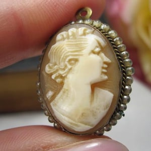 May include: An oval cameo pendant featuring a carved cream-colored profile of a person facing right, set against a translucent brown background. The piece is framed by a metallic border with a beaded edge and includes a small loop at the top.
