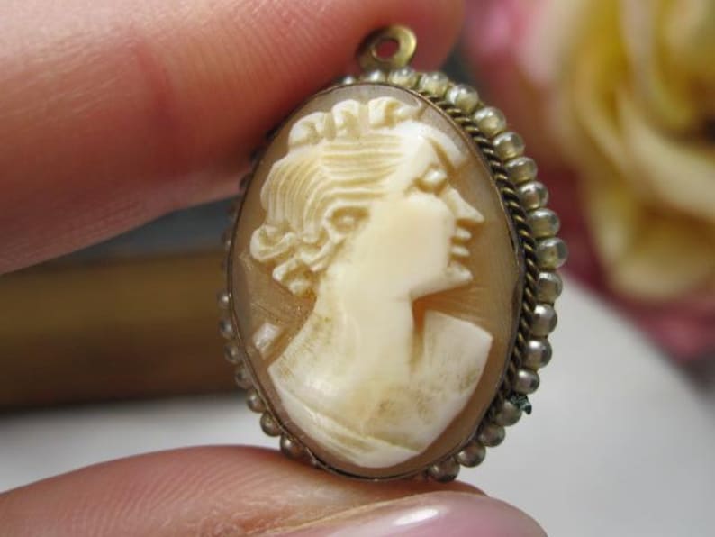May include: An oval cameo pendant featuring a carved cream-colored profile of a person facing right, set against a translucent brown background. The piece is framed by a metallic border with a beaded edge and includes a small loop at the top.
