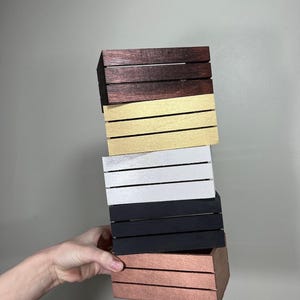 Könnte beinhalten: Ein Stapel von fünf kleinen Holzkisten in verschiedenen Farben: Dunkelbraun, Gold, Silber, Schwarz und Kupfer. Jede Kiste hat horizontale Lamellen. Die Kisten werden von einer Hand vor einem neutralen Hintergrund gehalten.