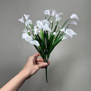 Könnte beinhalten: Ein Strauß weißer Calla-Lilien mit grünen Stielen und Blättern. Die Blumen werden in einer Hand vor einem neutralen grauen Hintergrund gehalten. Die Calla-Lilien haben ein glattes, elegantes Aussehen.