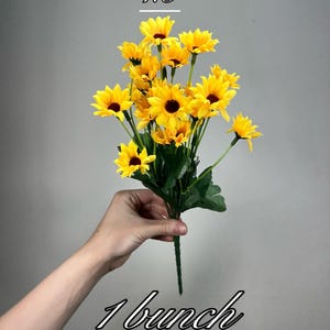 Puede incluir: Un ramo de girasoles artificiales de 15 pulgadas con pétalos amarillos y centros marrones. Las flores están dispuestas en un ramo y se sostienen en una mano. El texto "1 bunch" está escrito debajo del ramo.