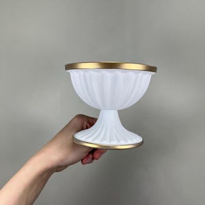 Op de afbeelding: Een witte, gegroefde glazen kom met een gouden rand en voet. De kom heeft een voetstuk en een decoratief, geribbeld ontwerp. De gouden rand voegt een vleugje elegantie toe aan het klassieke ontwerp. De kom wordt door een persoon vastgehouden.