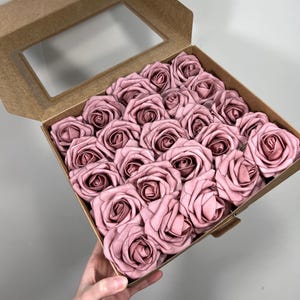 May include: A box of 25 dusty rose artificial roses. The roses are arranged inside a brown cardboard box with a clear window. The text "25 pcs" is written in cursive on the bottom right corner.
