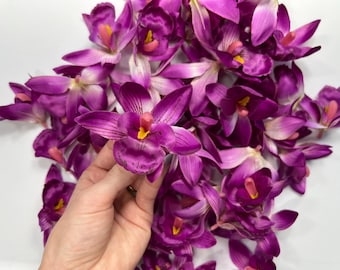 May include: A collection of artificial orchid flowers in vibrant shades of deep purple and magenta. Each flower features delicate, layered petals with subtle dark veining and a prominent yellow and pink center, creating a realistic floral display.