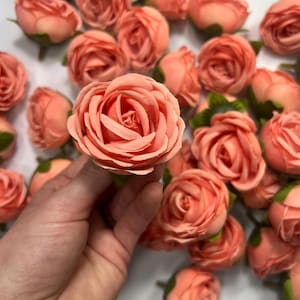 Può includere: Un mazzo di boccioli di rosa artificiali color pesca con steli verdi. I boccioli di rosa sono disposti in un mucchio su una superficie bianca. Una mano tiene uno dei boccioli di rosa.