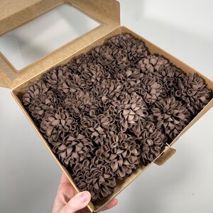 Pode incluir: Uma caixa de cartão com uma janela transparente contém numerosas flores de tecido castanho. As flores estão densamente embaladas, enchendo a caixa. A caixa é segurada, mostrando a cor e a textura das flores.