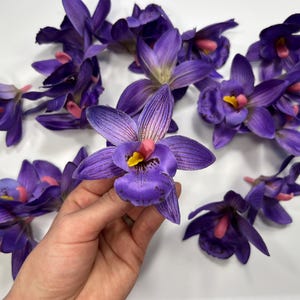 May include: A collection of purple and pink artificial orchid flowers. The flowers have a realistic appearance and are perfect for adding a touch of elegance to any space.