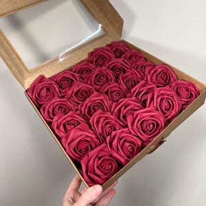 May include: A box of red roses. The roses are arranged in a square cardboard box with a clear window in the lid. The roses are a deep red color and appear to be made of fabric.