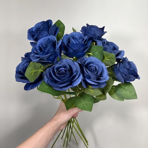 May include: A bouquet of deep blue roses with green leaves. The roses are in full bloom, and the stems are a light green color. The bouquet is held in a hand against a plain background.