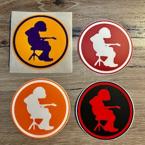 May include: Four round stickers with a silhouette of a person playing guitar. The stickers are in four different color combinations: yellow and purple, red and white, orange and white, and black and red.