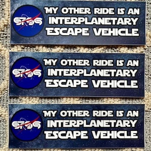 May include: Three blue and white stickers with a space-themed design. The stickers feature a stylized NASA logo and the text "MY OTHER RIDE IS AN INTERPLANETARY ESCAPE VEHICLE".