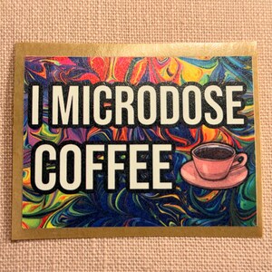 May include: A rectangular sticker with the text "I MICRODOSE COFFEE" in white letters with a black outline. The background is a colorful swirl of blues, greens, yellows, and reds. A pink coffee cup and saucer are on the right.
