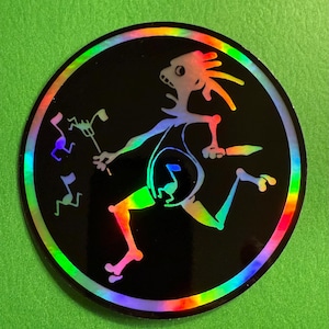 May include: A round, black sticker with a holographic rainbow border. The sticker features a stylized figure holding a musical instrument and running. Musical notes are floating nearby. The figure and notes have a rainbow gradient.