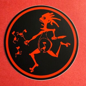 May include: A round sticker with a black background and a red border. The sticker features a red, stylized figure running with a musical note in its belly. Several smaller red figures are also present.