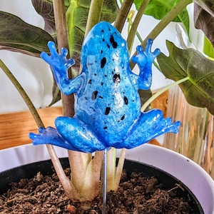 Puede incluir: Una estaca para plantas con forma de rana azul brillante con motas negras, colocada en una planta en maceta. La rana tiene los brazos y las piernas extendidos, añadiendo un toque caprichoso a la vegetación. La estaca es de metal.