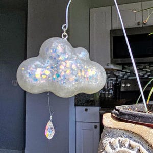 May include: A whimsical, cloud-shaped ornament with iridescent glitter embedded in a clear material. A crystal prism hangs below, suspended by a thin wire. The ornament is attached to a curved metal rod.